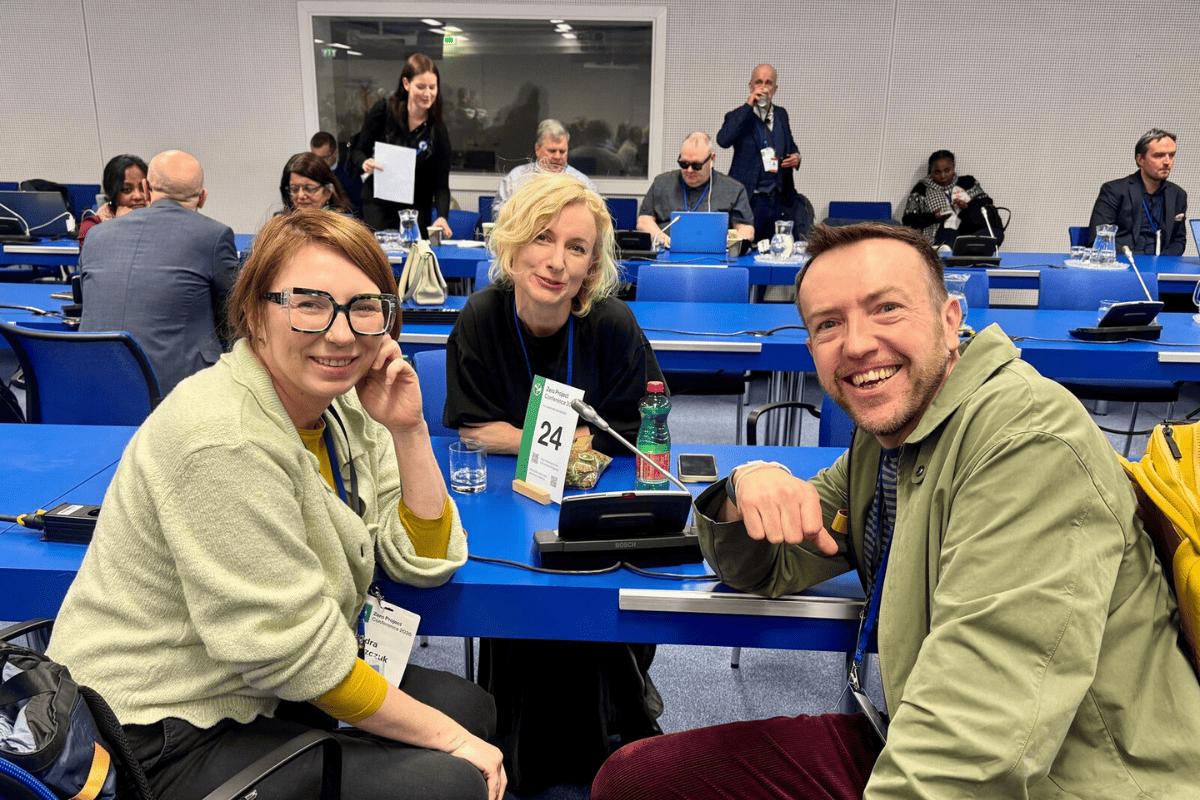 Trzy osoby siedzą przy stole konferencyjnym podczas Zero Project Conference. Na stole mikrofon i numer stanowiska 24. W tle inni uczestnicy przy niebieskich stołach.